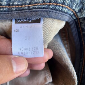 May include: Close-up of a denim jeans label with the brand name "Wrangler" in white. The label indicates a size of 30 inches. The jeans are a classic blue denim wash.