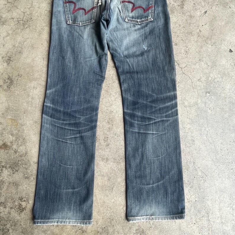 K&ouml;nnte beinhalten: Verblasste blaue Jeans mit geradem Bein. Die Jeans haben ein abgenutztes Aussehen mit sichtbaren Ausbleichungen und Abnutzungen. Die Ges&auml;&szlig;taschen haben ein markantes rotes Nahtdesign. Die Jeans liegen flach auf einer neutralen Oberfl&auml;che.