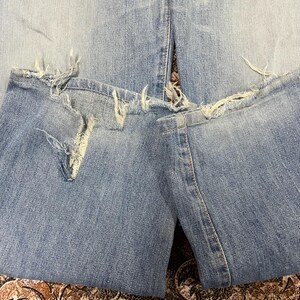 May include: Close-up of light blue denim jeans with distressed details. The jeans have ripped sections at the knees and frayed edges along the bottom hem, showcasing a worn, vintage aesthetic. The fabric appears faded.