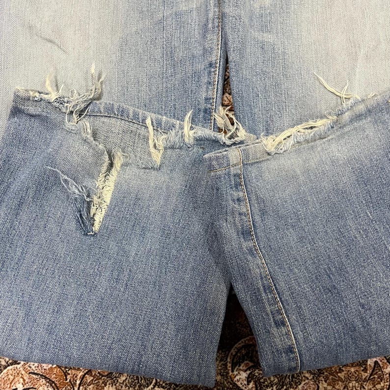 May include: Close-up of light blue denim jeans with distressed details. The jeans have ripped sections at the knees and frayed edges along the bottom hem, showcasing a worn, vintage aesthetic. The fabric appears faded.