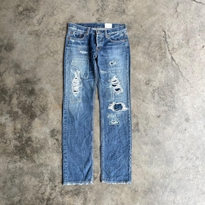 May include: Blue denim jeans with distressed details, including patches and ripped sections. The jeans have a button fly and a classic five-pocket design. The denim has a faded wash, giving it a worn appearance. The jeans are laid flat on a concrete surface.