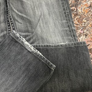 May include: Close-up of a pair of faded black denim jeans. The jeans are folded, showing the hem and the texture of the fabric. The stitching and details of the denim are visible.