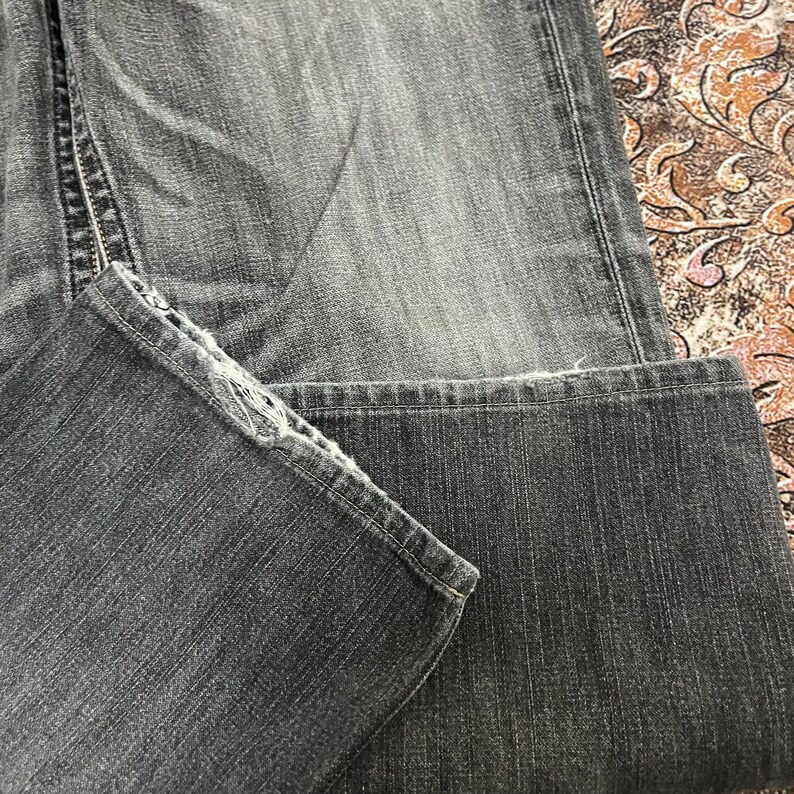 May include: Close-up of a pair of faded black denim jeans. The jeans are folded, showing the hem and the texture of the fabric. The stitching and details of the denim are visible.