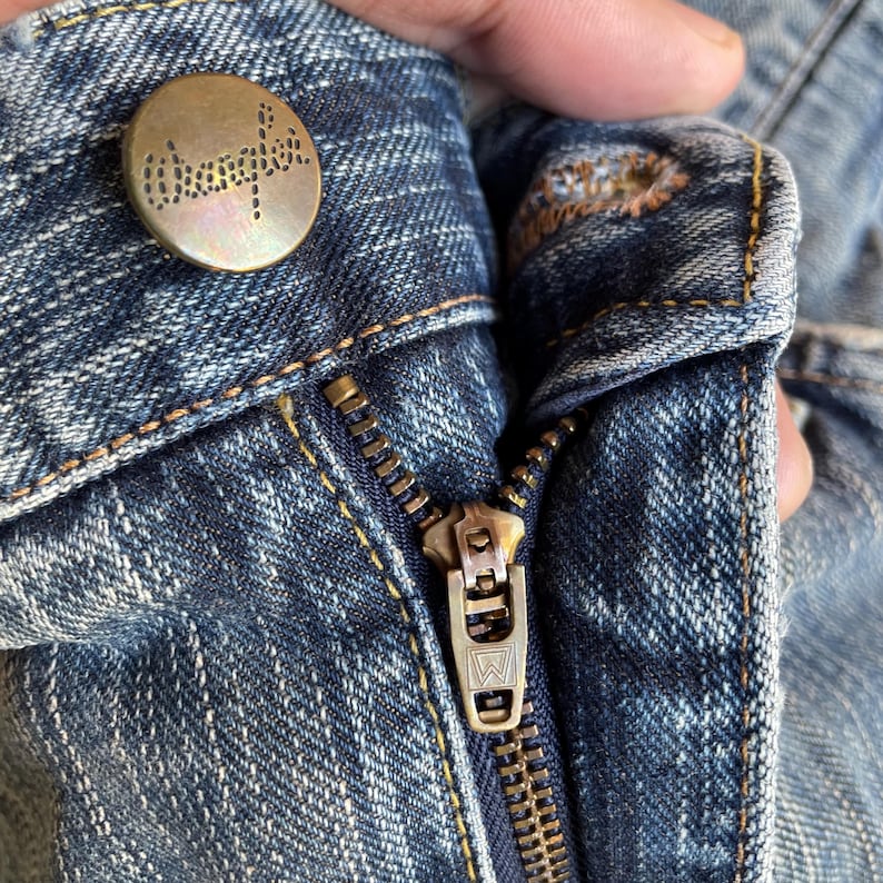 May include: Close-up of blue denim jeans with a brass-colored button and zipper. The button is embossed with the brand name. The zipper is partially open, revealing the metal teeth and pull tab. The denim has a worn appearance.