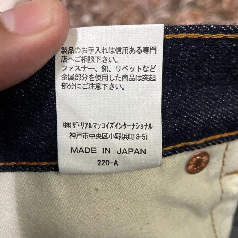May include: Close-up of a tag on denim jeans. The tag has Japanese text and the words "MADE IN JAPAN". The tag also includes the number "220-A". A copper-colored button is visible.