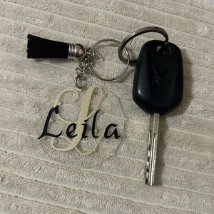 Peut inclure: Un porte-cl&eacute;s personnalis&eacute; avec le nom "Leila" en &eacute;criture noire sur un cercle acrylique transparent, avec une initiale dor&eacute;e paillet&eacute;e. Comprend un pompon noir et une cl&eacute; de voiture.