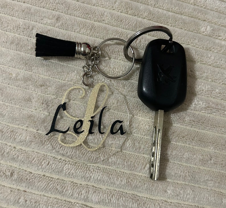 Peut inclure: Un porte-cl&eacute;s personnalis&eacute; avec le nom "Leila" en &eacute;criture noire sur un cercle acrylique transparent, avec une initiale dor&eacute;e paillet&eacute;e. Comprend un pompon noir et une cl&eacute; de voiture.