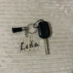 Peut inclure: Une cl&eacute; de voiture avec un bo&icirc;tier noir et une cl&eacute; argent&eacute;e, attach&eacute;e &agrave; un porte-cl&eacute;s avec un pompon noir et un charm en acrylique transparent. Le charm porte le nom "Leila" en &eacute;criture cursive.