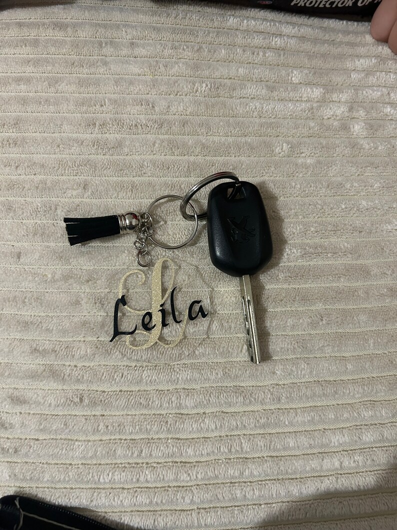 Peut inclure: Une cl&eacute; de voiture avec un bo&icirc;tier noir et une cl&eacute; argent&eacute;e, attach&eacute;e &agrave; un porte-cl&eacute;s avec un pompon noir et un charm en acrylique transparent. Le charm porte le nom "Leila" en &eacute;criture cursive.