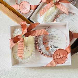 May include: White gift boxes with coral ribbon and embellishments. Each box contains a pearl strand, a decorative item with a crystal-like appearance, and a card with Arabic text. The boxes are sealed with a round sticker that says "Elena".