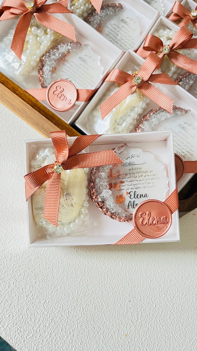 May include: White gift boxes with coral ribbon and embellishments. Each box contains a pearl strand, a decorative item with a crystal-like appearance, and a card with Arabic text. The boxes are sealed with a round sticker that says "Elena".