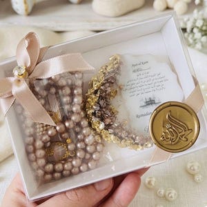 May include: A clear box contains a pearl necklace, a gold-colored crescent moon decoration, and a card with Arabic text. The box is tied with a pink ribbon and a gold-colored jewel. The box is decorated with a gold-colored seal.