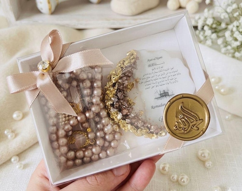 May include: A clear box contains a pearl necklace, a gold-colored crescent moon decoration, and a card with Arabic text. The box is tied with a pink ribbon and a gold-colored jewel. The box is decorated with a gold-colored seal.