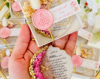 May include: A pink and gold resin ornament with an Arabic inscription. The ornament is shaped like a crescent moon and has a gold chain attached to it. The inscription reads "Allah la ilaha illa huwa al-hay al-qayyum" which translates to "Allah, there is no god but He, the Ever-Living, the Self-Sustaining."