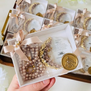 May include: A collection of gift boxes, each containing a pearl rosary and a crescent moon decoration. The boxes are tied with light pink ribbons and adorned with a gold seal. The theme appears to be related to Ramadan.
