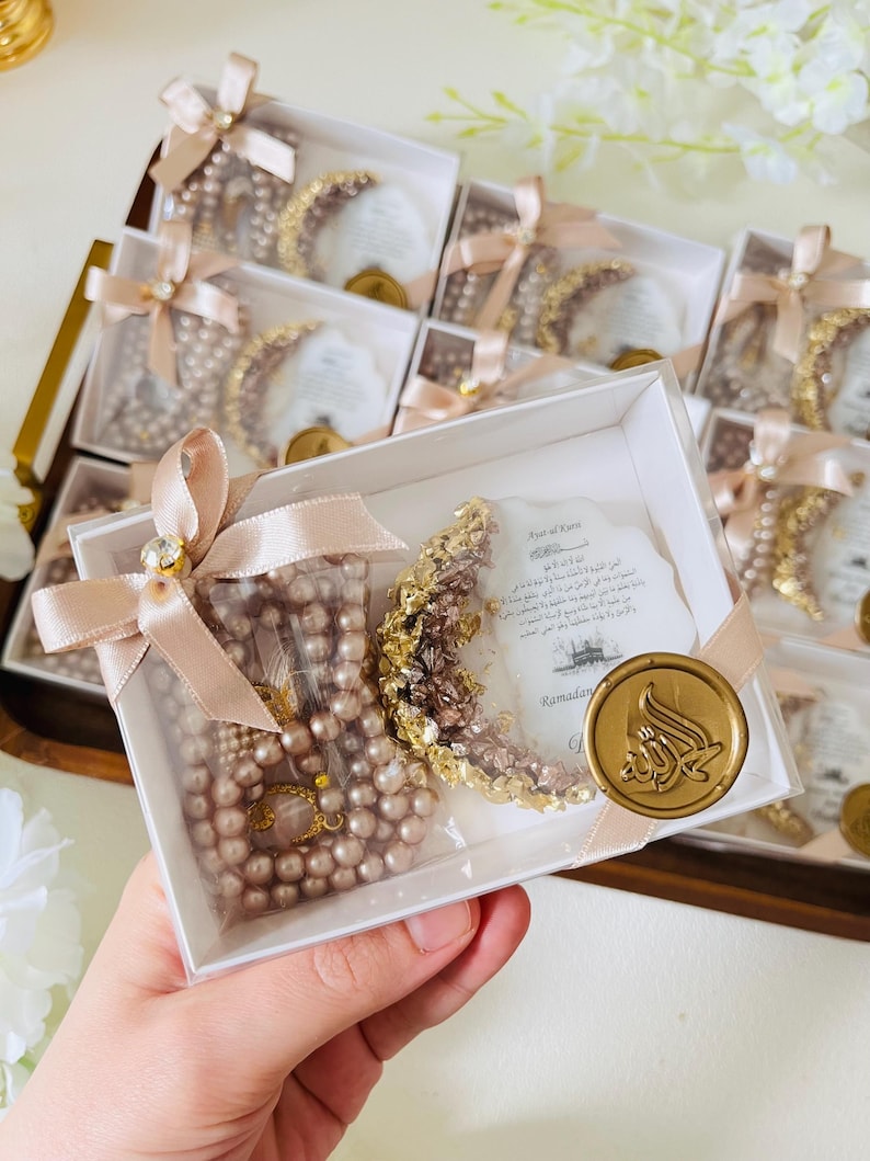 May include: A collection of gift boxes, each containing a pearl rosary and a crescent moon decoration. The boxes are tied with light pink ribbons and adorned with a gold seal. The theme appears to be related to Ramadan.