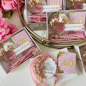 May include: Pink and gold prayer beads with a pink velvet prayer book and a white ceramic tile with an Islamic prayer in Arabic script. The tile has a gold crescent moon and crystals embedded in it. The prayer beads have a pink tassel.