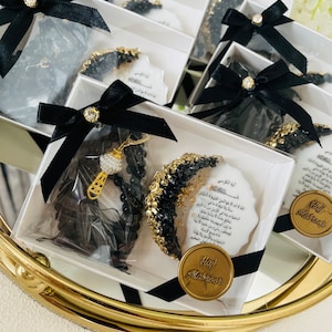 May include: Several gift boxes with black bows and gold accents are arranged on a gold-rimmed tray. Each box contains a black beaded necklace, a decorative item with gold and black embellishments, and a card with Arabic text. The boxes are tied with black ribbons.