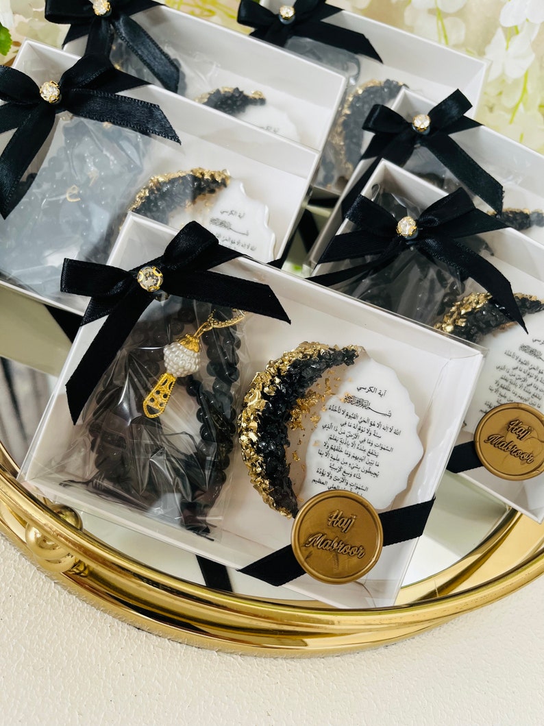May include: Several gift boxes with black bows and gold accents are arranged on a gold-rimmed tray. Each box contains a black beaded necklace, a decorative item with gold and black embellishments, and a card with Arabic text. The boxes are tied with black ribbons.