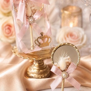 Könnte beinhalten: Ein runder Handspiegel mit goldfarbenem Rahmen und Griff, verziert mit einer rosa Satinschleife, einer zentralen Kunstperle und getrockneten beigen Blumen. Ein zweiter identischer Spiegel befindet sich in einer transparenten Box mit Kronenanhänger.