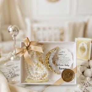May include: A white gift box tied with a gold ribbon, containing a pearl necklace, a decorative crescent, and a card with text. The crescent is gold and crystal. The card says "Welcome Baby Karam". A gold seal is on the box.