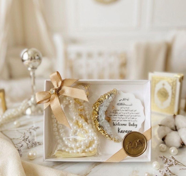 May include: A white gift box tied with a gold ribbon, containing a pearl necklace, a decorative crescent, and a card with text. The crescent is gold and crystal. The card says "Welcome Baby Karam". A gold seal is on the box.