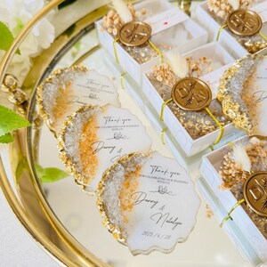 May include: Gold and white wedding favors with a wax seal and dried flowers. The favors are in small boxes and have a thank you message for celebrating the wedding with the couple, Danny and Natalya, and the date 2025/6/28.