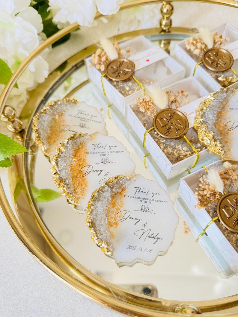 May include: Gold and white wedding favors with a wax seal and dried flowers. The favors are in small boxes and have a thank you message for celebrating the wedding with the couple, Danny and Natalya, and the date 2025/6/28.