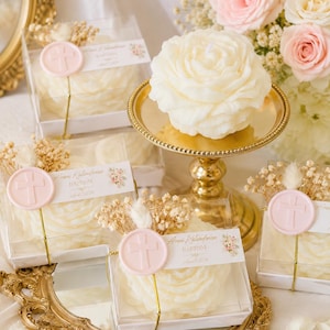 May include: Cream-colored rose-shaped candles in clear square boxes. Each box features a pink wax seal with a cross, a sprig of dried baby's breath, and a white label printed with the text Arni Kalandarian BAPTISM and a date in April 2024.