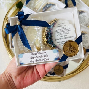May include: A clear box containing a pearl strand, a crescent-shaped decoration with blue and gold accents, and a card with Arabic text. The box is tied with a navy blue ribbon and a gold seal. The text reads "Proud Parents Rezaul & Rusaylee Big Brother Rayn."