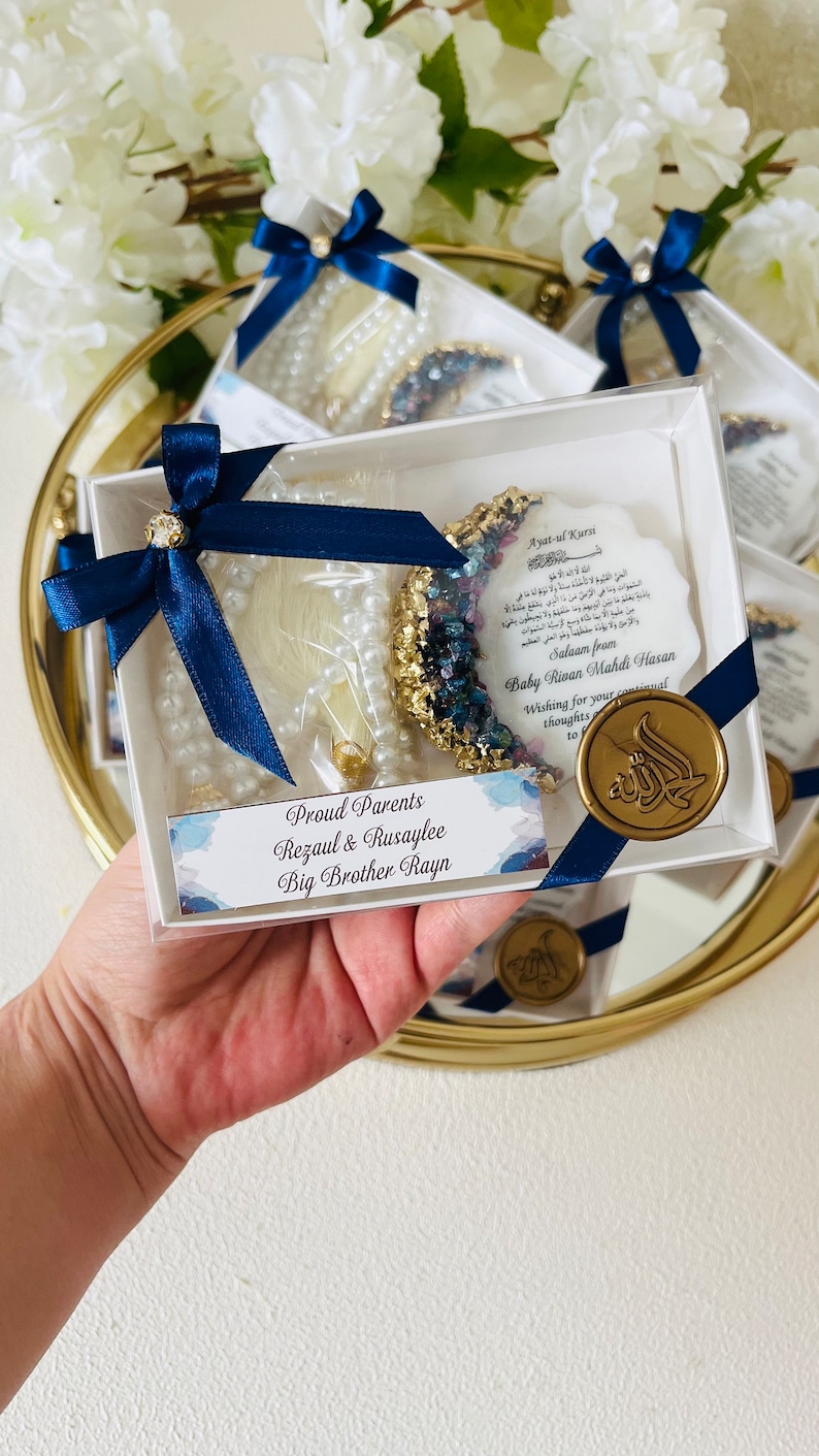 May include: A clear box containing a pearl strand, a crescent-shaped decoration with blue and gold accents, and a card with Arabic text. The box is tied with a navy blue ribbon and a gold seal. The text reads "Proud Parents Rezaul & Rusaylee Big Brother Rayn."
