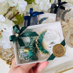 May include: A gift box with a green beaded prayer beads, a crescent moon decoration, and a gold-colored seal. The box is tied with a green ribbon. The background includes white flowers and other gift boxes.