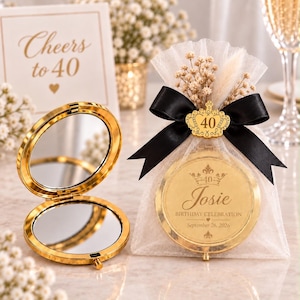 Pode incluir: Um espelho de bolso circular dourado com tampa aberta e um segundo espelho dentro de um saco de tule branco. O saco apresenta uma fita de cetim preta, um pendente de coroa dourada com o número 40 e o texto Josie Birthday Celebration.