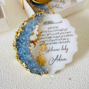 May include: A white, round, resin ornament with a blue and gold glitter design. The ornament features an Arabic inscription and the text "Welcome baby Adam".