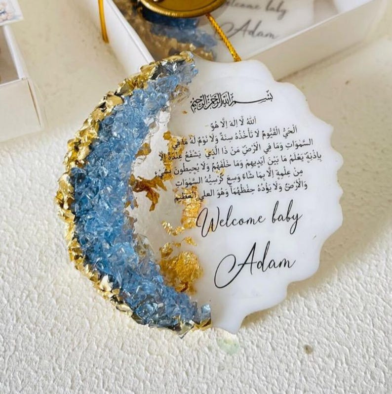 May include: A white, round, resin ornament with a blue and gold glitter design. The ornament features an Arabic inscription and the text "Welcome baby Adam".