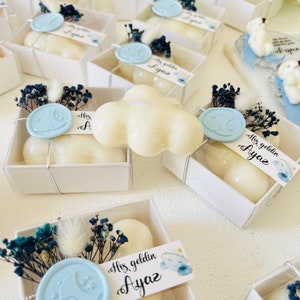 May include: White favour boxes with blue and white floral accents, filled with white scented candles. The boxes have a blue wax seal with a crescent moon and cloud design. The boxes have a label that says "Hoş geldin Ayaz" which translates to "Welcome Ayaz".