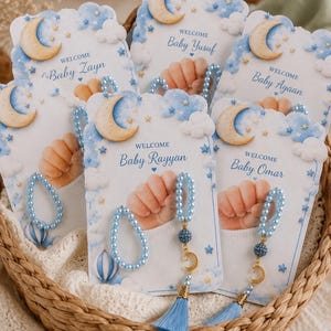 May include: A collection of baby shower favor cards featuring a blue sky, clouds, stars, and a crescent moon. Each card displays a baby's hand and the text Welcome Baby followed by names like Zayn, Yusuf, Ayaan, Rayyan, and Omar. Attached are light blue pearl bead bracelets and charms with tassels.