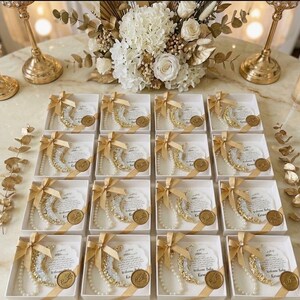 May include: Arrangement of white gift boxes, each containing a crescent moon-shaped decoration with gold and pearl accents, tied with a gold ribbon. A gold medallion and a card with text are also included. The boxes are arranged on a marble surface.