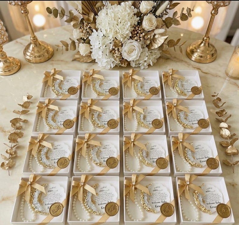 May include: Arrangement of white gift boxes, each containing a crescent moon-shaped decoration with gold and pearl accents, tied with a gold ribbon. A gold medallion and a card with text are also included. The boxes are arranged on a marble surface.