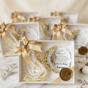 May include: White gift boxes tied with gold ribbons, each containing a pearl necklace, a crescent-shaped decoration, and a card with Arabic text. The boxes are decorated with gold accents and a wax seal.