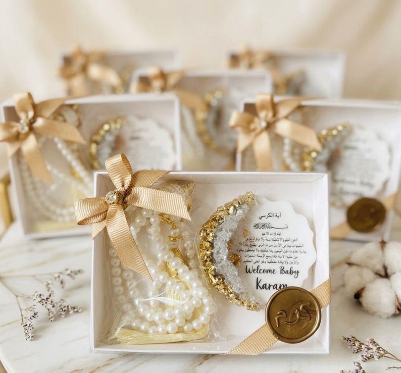 May include: White gift boxes tied with gold ribbons, each containing a pearl necklace, a crescent-shaped decoration, and a card with Arabic text. The boxes are decorated with gold accents and a wax seal.