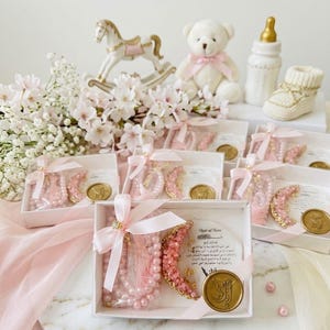 May include: A collection of baby shower favors, including pink pearl bracelets, a teddy bear, a rocking horse, and a baby bottle. The favors are presented in white boxes with pink ribbons and gold accents, creating a soft and elegant aesthetic.