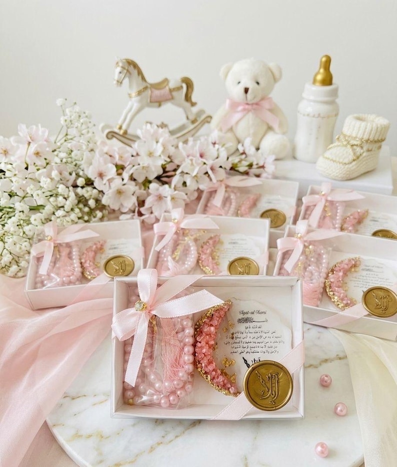 May include: A collection of baby shower favors, including pink pearl bracelets, a teddy bear, a rocking horse, and a baby bottle. The favors are presented in white boxes with pink ribbons and gold accents, creating a soft and elegant aesthetic.