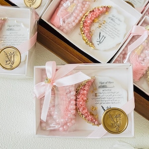 May include: Pink prayer beads and a crescent-shaped decoration with gold accents are presented in white boxes, tied with pink ribbons. Each box includes a gold-colored seal with Arabic script and a card with Arabic text.