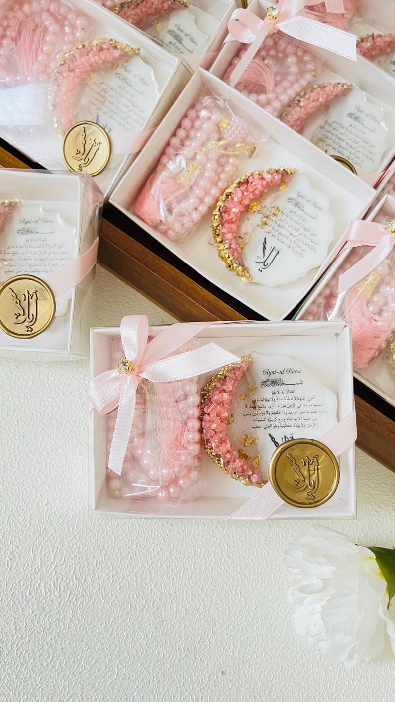 May include: Pink prayer beads and a crescent-shaped decoration with gold accents are presented in white boxes, tied with pink ribbons. Each box includes a gold-colored seal with Arabic script and a card with Arabic text.