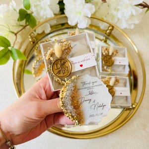 May include: White and gold wedding favor boxes with a clear top. Each box contains a small white and gold resin coaster with a gold wax seal and a thank you message. The message reads "Thank you for celebrating our wedding with us, Danny & Natalya, 2025/6/28".