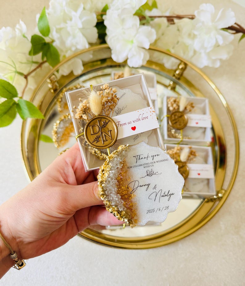 May include: White and gold wedding favor boxes with a clear top. Each box contains a small white and gold resin coaster with a gold wax seal and a thank you message. The message reads "Thank you for celebrating our wedding with us, Danny & Natalya, 2025/6/28".