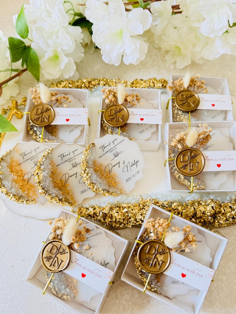 May include: Gold and white wedding favor boxes with a wax seal and dried flowers. The boxes have a thank you message and the date of the wedding. The text reads "Thank you for celebrating our wedding with us. Danny & Natalie 2023/1/28. From us with love."