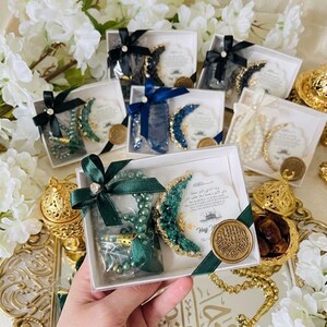 May include: Gift boxes with green, blue, and white prayer beads, crescent moon decorations, and cards. Each box is tied with a ribbon. Gold-colored decorative items and white flowers are in the background.