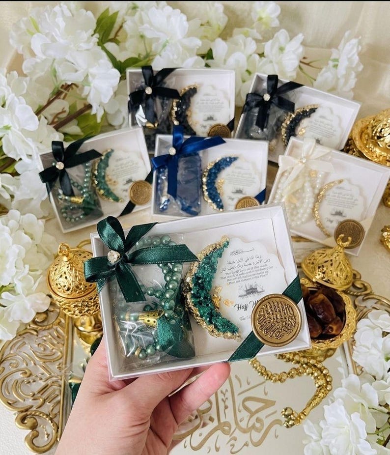 May include: Gift boxes with green, blue, and white prayer beads, crescent moon decorations, and cards. Each box is tied with a ribbon. Gold-colored decorative items and white flowers are in the background.